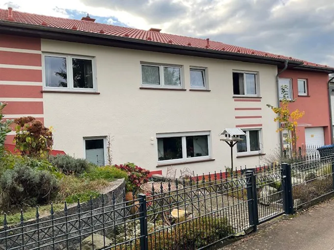 Zweistöckiges Wohnhaus mit rotem Dach, heller, zweifarbiger Fassade und mehreren Fenstern, davor ein kleiner Vorgarten mit Zaun.
